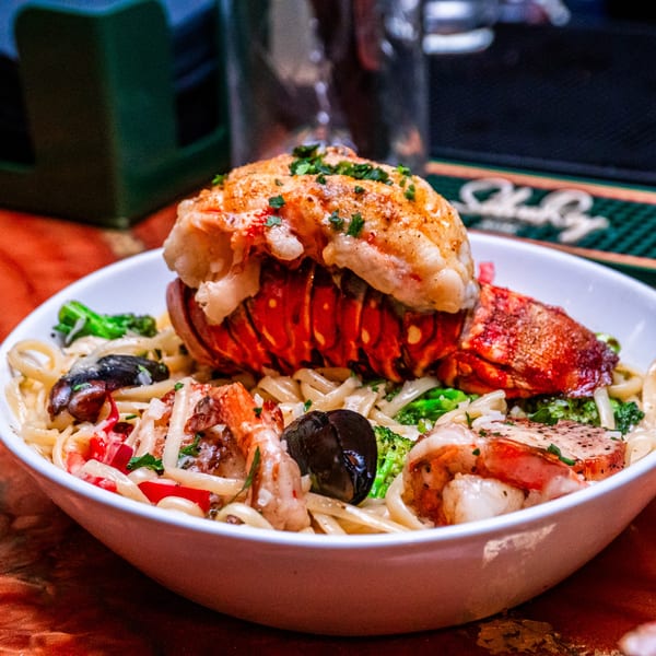 Lobster tail and shrimp pasta with broccoli and mushrooms in a white bowl.