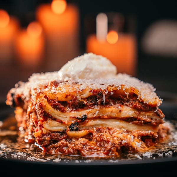 A close-up of a layered lasagna with melted cheese and a dollop of white cream, against a blurred background of warm candlelight.